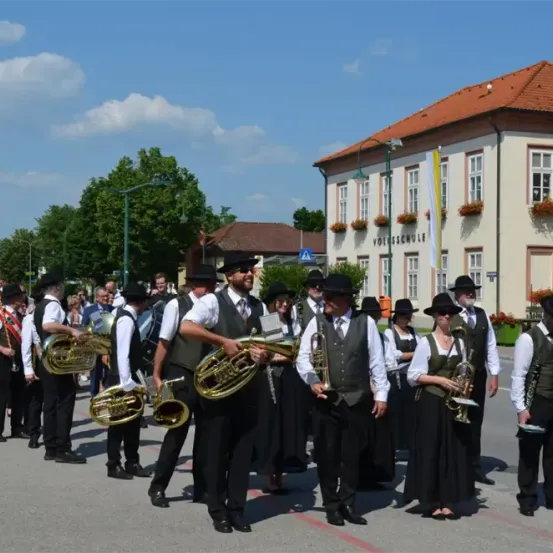 Eine Blaskapelle steht vor einem Gebäude mit einem roten Dach. Die Bandmitglieder tragen schwarze Hüte, Westen und halten Blechblasinstrumente. Bäume und Straßenlaternen sind im Hintergrund zu sehen.