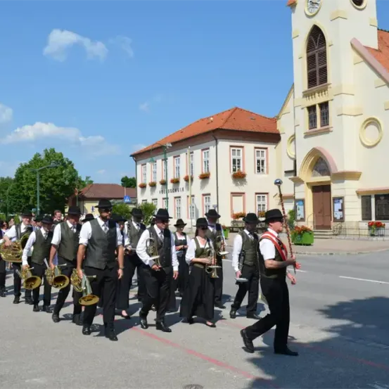 Eine Blaskapelle in traditioneller Kleidung tritt vor einer historischen Kirche an einem sonnigen Tag auf.