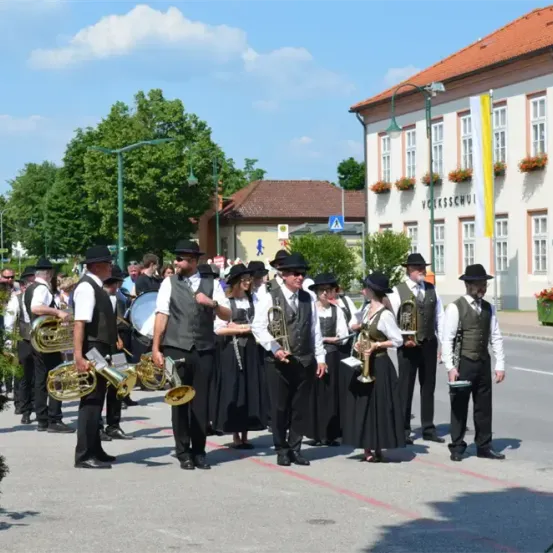 Ein Blasorchester in traditioneller Kleidung spielt auf der Straße, mit einem Gebäude, das eine Flagge und Topfpflanzen im Hintergrund zeigt.