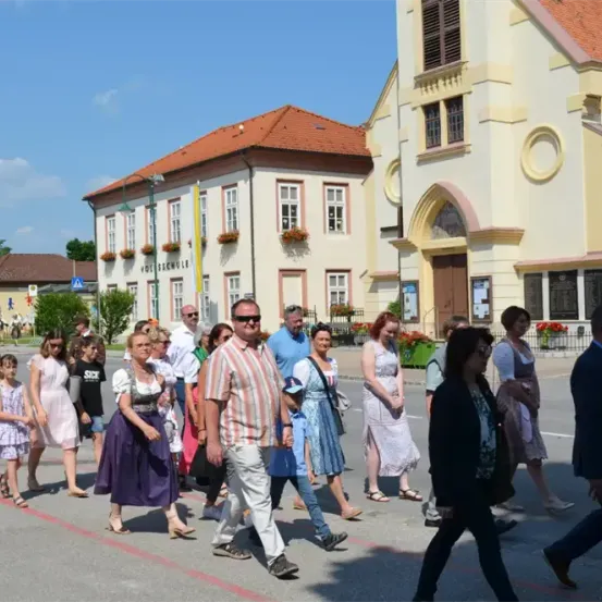 Eine Gruppe von Menschen, darunter Erwachsene und Kinder, geht in einer Reihe auf einer Straße vor einem Kirchengebäude. Das Gebäude hat ein rotes Dach und mehrere Fenster. Einige Leute tragen Brillen und Schuhe, während andere Hüte tragen.