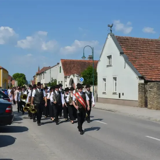 Eine Gruppe von Menschen in schwarzen Outfits und Hüten marschiert auf der Straße und spielt Musikinstrumente. Die Straße hat geparkte Autos auf beiden Seiten. Gebäude mit Ziegeldächern befinden sich an den Seiten.