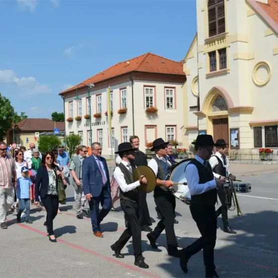 Eine Gruppe von Menschen, darunter Musiker mit Trommeln und Trompeten, geht an einer Kirche an einem sonnigen Tag vorbei.