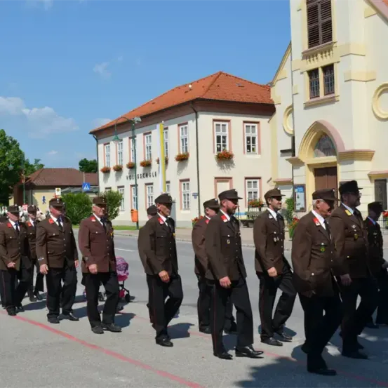 Eine Gruppe uniformierter Männer, möglicherweise Militärangehörige, marschiert auf einer Straße. Im Hintergrund befinden sich Gebäude mit roten Dächern und weißen Wänden.
