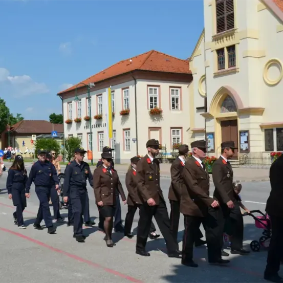 Eine Gruppe von Menschen in Uniform marschiert vor einem Gebäude mit rotem Dach und großem Bogen die Straße entlang.
