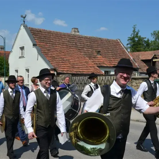 Eine Blaskapelle, gekleidet in traditioneller Kleidung, zieht durch ein Dorf, wobei die Leute zuschauen und Instrumente wie Trompete und Trommeln spielen.
