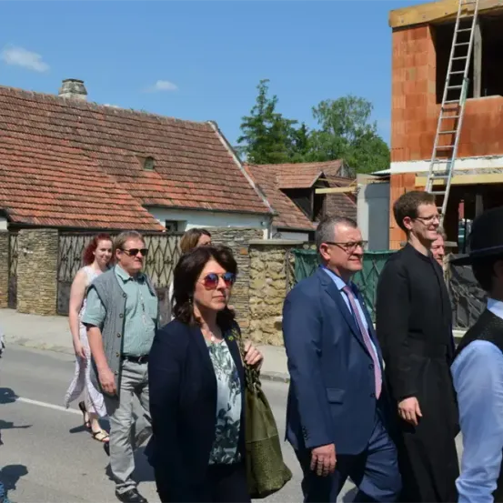 Eine Gruppe von Menschen, einschließlich Erwachsener und eines Kindes, gehen auf einer Straße. Einige tragen Sonnenbrillen, während andere formell gekleidet sind. Sie scheinen in ein Gespräch vertieft zu sein. Im Hintergrund befindet sich ein Haus im Bau.