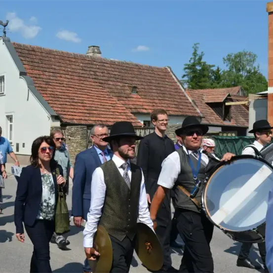 Eine Gruppe von Menschen geht in einem Dorf mit traditioneller Kleidung, einige tragen Hüte und halten Musikinstrumente wie Trommeln und Becken, mit einem Haus und Bäumen im Hintergrund.