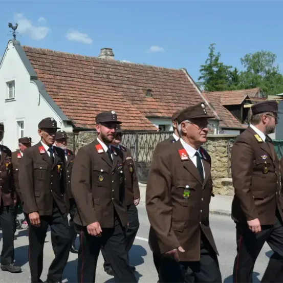 Eine Gruppe uniformierter Männer marschiert bei einer Parade, trägt Hüte, Medaillen und schwarze Krawatten. Sie gehen auf einer Straße vor traditionellen Häusern mit roten Dächern.