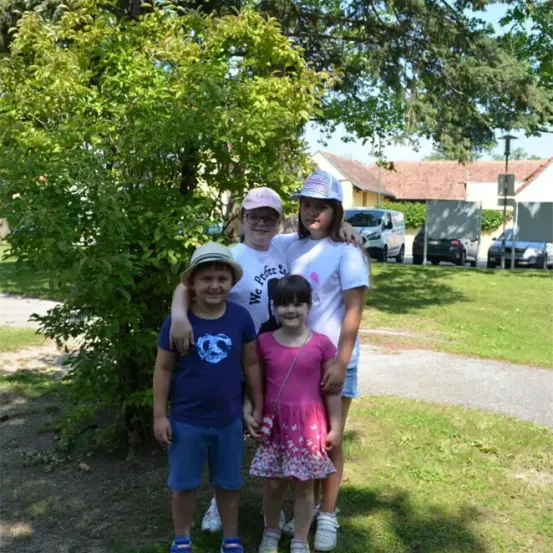 Vier Kinder stehen vor einem Baum, zwei Mädchen in Pink und Weiß, ein Junge in Blau und ein weiterer Junge in Weiß. Dahinter ist ein Haus mit einem braunen Dach zu sehen.