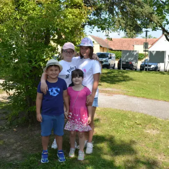 Vier Kinder stehen in einem Park mit einem Baum, zwei Mädchen tragen Hüte, eines mit Brille, eines in einem rosa Kleid. Dahinter sind Autos geparkt und ein Haus.