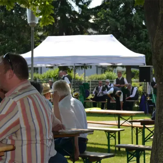 Men sitzen unter einem Zelt in einem Park an Tischen, mit Musikern im Hintergrund und Bäumen in der Ferne.