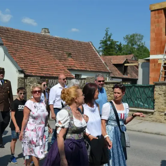 Eine Gruppe von Menschen, einige in traditioneller Kleidung, geht auf einer Straße vor Häusern. Ein Mann in Uniform steht in der Nähe.