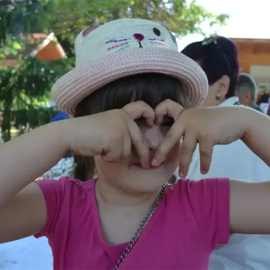 Ein kleines Mädchen trägt einen rosafarbenen Katzenhut und formt mit ihren Händen ein Herz vor ihrem Gesicht. Sie trägt ein rosa T-Shirt und eine Halskette. Im Hintergrund stehen mehrere Personen, vermutlich bei einer Veranstaltung.