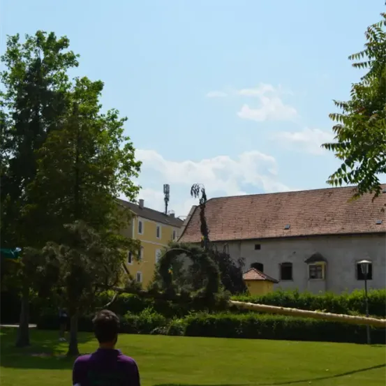 Ein Mann steht in einem Grasbereich vor einem Gebäude mit gelber Fassade und braunem Dach. Bäume umgeben den Bereich, und im Hintergrund befindet sich eine hohe Struktur.