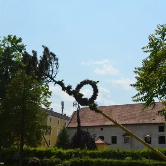 Ein Haus mit einem braunen Dach und gelben Wänden steht inmitten üppiger Grünflächen. Ein Baumzweig, geschmückt mit einem Kranz, erstreckt sich in den Himmel. Der Himmel ist blau mit verstreuten Wolken.