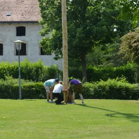 Drei Männer arbeiten an einem Pfahl auf einer grasigen Wiese. Sie tragen Mützen und Turnschuhe. Ein großes weißes Gebäude ist im Hintergrund.