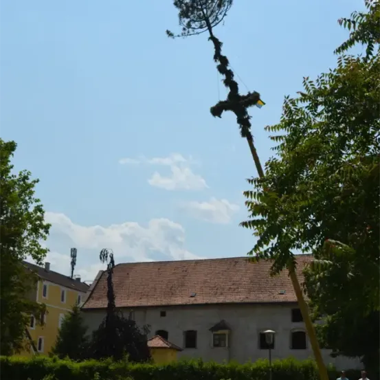 Ein Baum mit einer gelben Flagge wird über einem Gebäude mit braunem Dach und gelben Wänden unter einem bewölkten Himmel gehoben. Menschen gehen vor dem Gebäude.
