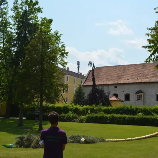 Eine Person in einem lila Shirt steht auf einem Rasen. Hinter ihr befindet sich ein großes Gebäude mit einem braunen Dach. Vor dem Gebäude stehen Büsche und Bäume. Eine Person geht im Hintergrund.