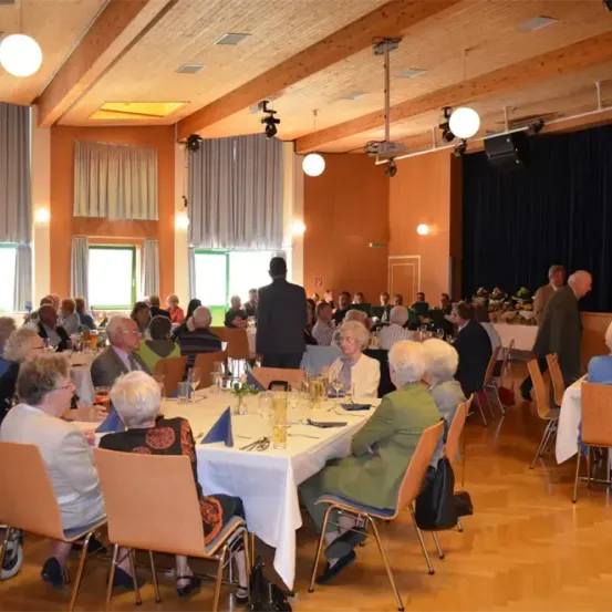 Eine große Gruppe von Menschen, überwiegend Senioren, ist in einem Bankettsaal versammelt. Sie sitzen um Tische mit weißen Tischdecken, auf denen verschiedene Gegenstände stehen. Der Raum ist gut beleuchtet und hat Holzböden.