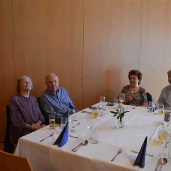 Vier ältere Personen sitzen um einen Tisch herum, wahrscheinlich in einem Restaurant. Der Tisch ist mit Weingläsern, Tellern, Gabeln und einer Vase mit Blumen gedeckt. Der Raum hat Holzwände.