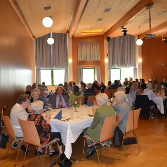 Ein Speisesaal mit mehreren Tischen, an denen Personen sitzen. Einige Tische haben Blumen und Gläser, und der Raum hat braune Wände und Holzböden. Lampen hängen von der Decke, und Fenster mit Vorhängen sind sichtbar.