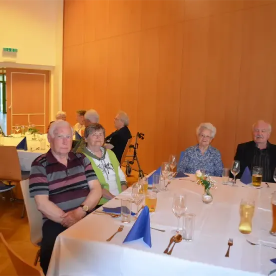 Eine Gruppe älterer Personen sitzt an einem langen Tisch mit Weingläsern, Bierkrügen und Servietten. Sie befinden sich in einem Essbereich mit Holzböden und orangefarbenen Wänden.