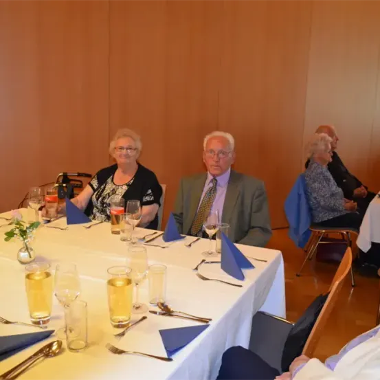 Mehrere Menschen sitzen an einem Tisch mit Weingläsern und Tellern. Eine ältere Frau trägt eine Brille und eine Halskette. Ein älterer Mann trägt eine Krawatte. Der Tisch ist mit Blumen, Besteck und blauen Servietten gedeckt.