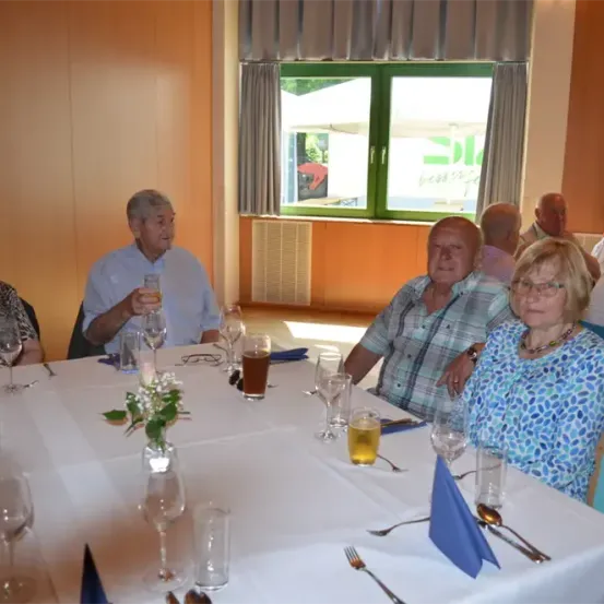 Eine Gruppe älterer Menschen sitzt um einen Tisch mit Wein glasses, einer Blume und Besteck herum. Sie nehmen wahrscheinlich an einer Versammlung oder einem Treffen teil.