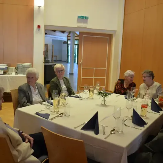 Eine Gruppe älterer Frauen sitzt um einen Tisch mit Weingläsern und Tellern herum, eine hält ein Glas Bier. Im Hintergrund ist eine Tür geöffnet.