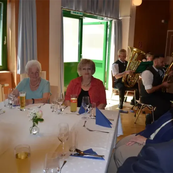 Eine Innenszene beim Essen mit zwei Frauen am Tisch, Weingläsern, einer Blume und einem Tubaspieler im Hintergrund.