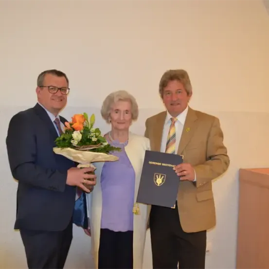 Drei Menschen stehen für ein Foto zusammen. Eine Frau in der Mitte hält einen Blumenstrauß und ein Zertifikat, während die beiden Männer Zertifikate halten. Sie alle lächeln.