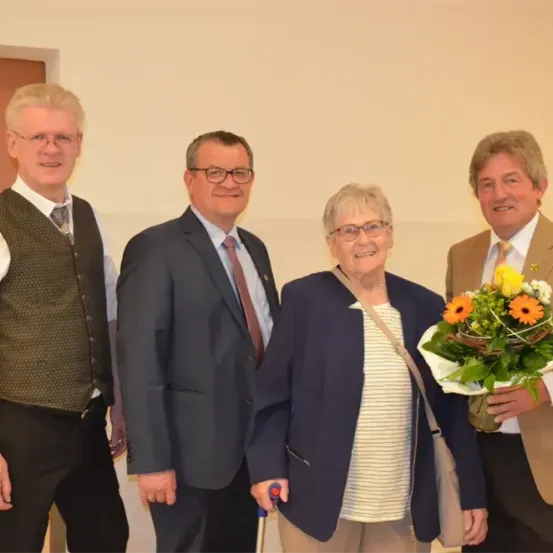 Vier Personen stehen in einer Reihe und lächeln für ein Foto. Eine Frau in der Mitte hält einen Blumenstrauß. Die Personen sind formell gekleidet.