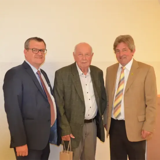 Drei Männer in Anzügen und Krawatten stehen zusammen. Der Mann auf der linken Seite trägt eine Brille. Der Mann in der Mitte hält einen Papierbeutel. Der Mann auf der rechten Seite hat eine gestreifte Krawatte.