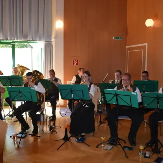 Ein Blasorchester mit Tuba- und Trompetenspielern tritt in einem Raum mit Holzboden und braunen Wänden auf.