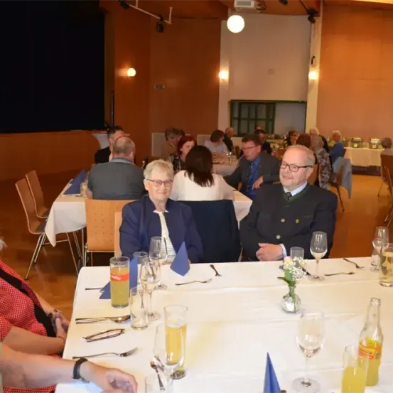 Eine Gruppe von Menschen sitzt in einem Speisesaal um einen Tisch herum und trägt formelle Kleidung. Der Tisch ist mit Weingläsern, einer Blumenvase und einer Flasche gedeckt. Der Raum hat Stühle, Tische und eine Projektionsleinwand. Die Wände sind in einer warmen Farbe gestrichen, und es sind Lampen an ihnen montiert.