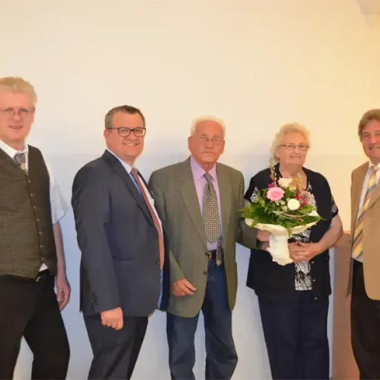 Fünf Personen, feierlich gekleidet, stehen gemeinsam vor einem weißen Hintergrund. Die zentrale Figur hält einen Blumenstrauß.