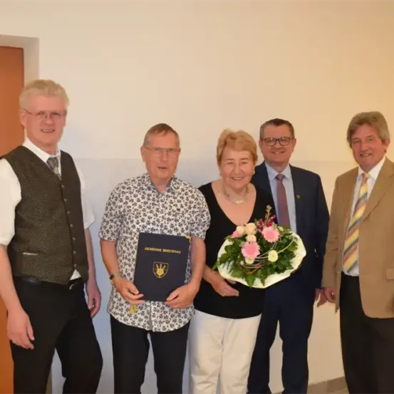 Fünf Menschen stehen lächelnd in einem Raum. Ein Mann in der Mitte hält ein Buch und einen Blumenstrauß. Eine weitere Frau hält einen Blumenstrauß. Hinter ihnen ist eine weiße Wand sichtbar.