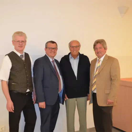 Vier Männer in Anzügen und Krawatten stehen gegen eine helle Wand. Sie scheinen für ein Foto zu posieren.