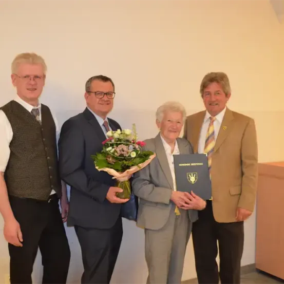 Vier Personen stehen zusammen und lächeln. Der Mann auf der linken Seite trägt eine Weste und ein weißes Hemd. Der Mann in der Mitte hält einen Blumenstrauß und eine blaue Mappe. Die Frau in der Mitte trägt einen grauen Anzug und hält eine Mappe. Der Mann auf der rechten Seite trägt einen braunen Anzug und eine Krawatte.