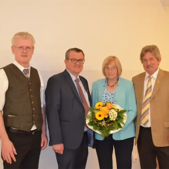 Vier Menschen stehen in einem Raum zusammen. Eine Frau hält einen Blumenstrauß. Die anderen tragen Anzüge und Krawatten.