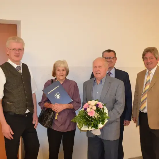 Fünf Menschen stehen lächelnd in einem Raum. Eine ältere Frau hält einen Blumenstrauß und ein Buch, während ein älterer Mann ein Buch hält. Hinter ihnen sehen zwei Männer in Anzügen zu.