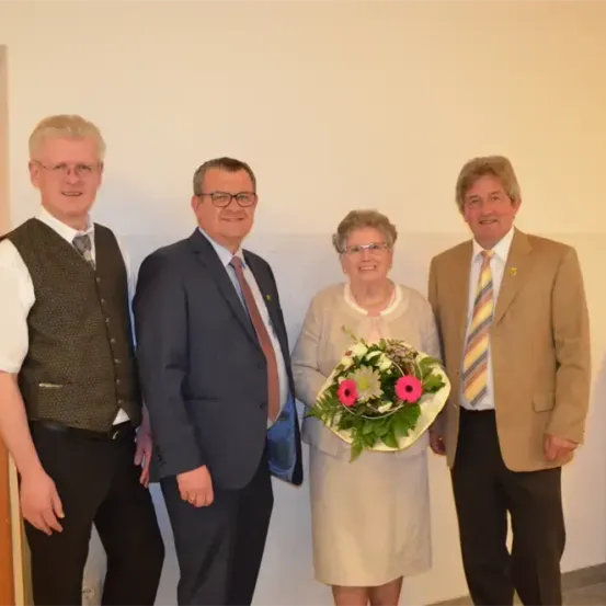 Vier Menschen stehen lächelnd in einem Raum zusammen. Eine ältere Frau hält einen Blumenstrauß. Der Mann links trägt eine Weste und eine Brille. Die Männer in der Mitte und rechts tragen Anzüge und Krawatten.