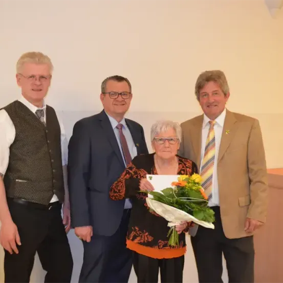 Eine Frau hält einen Blumenstrauß und ein Zertifikat in der Hand, lächelt und wird von drei Männern in formeller Kleidung umgeben. Sie stehen vor einer weißen Wand.