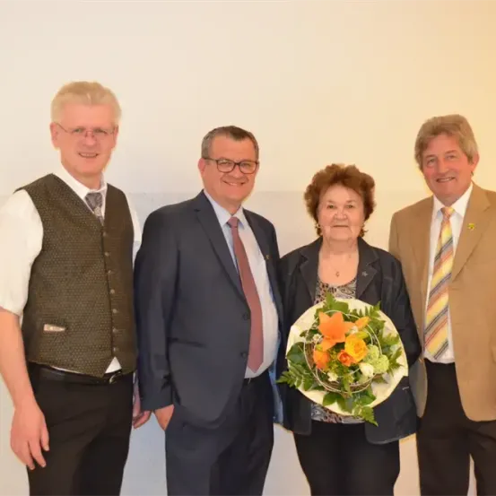 Vier Personen stehen vor einer weißen Wand. Die Frau hält einen Blumenstrauß, während die Männer Anzüge und Krawatten tragen.