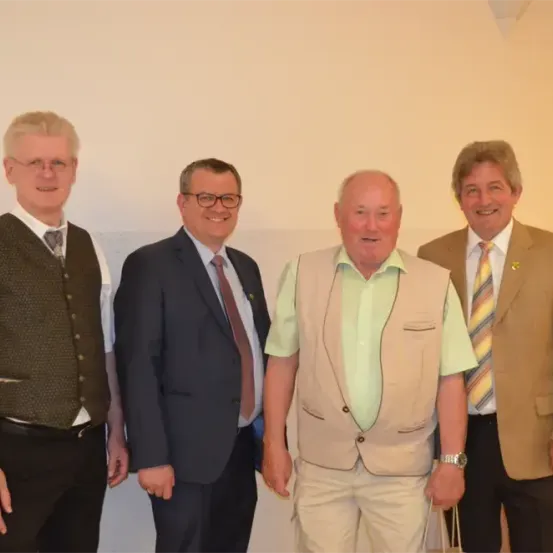 Vier Männer in einer Reihe stehend, alle lächelnd, einer hält eine Tasche, in einem hellen Raum.