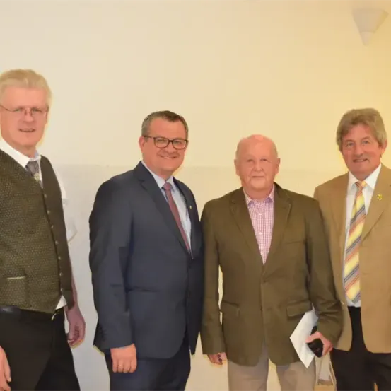 Vier Männer stehen zusammen, wahrscheinlich in einer professionellen Umgebung. Der Mann auf der linken Seite trägt eine Weste, während der Mann in der Mitte einen Anzug und eine Krawatte trägt. Der Mann auf der rechten Seite hält ein Papier.