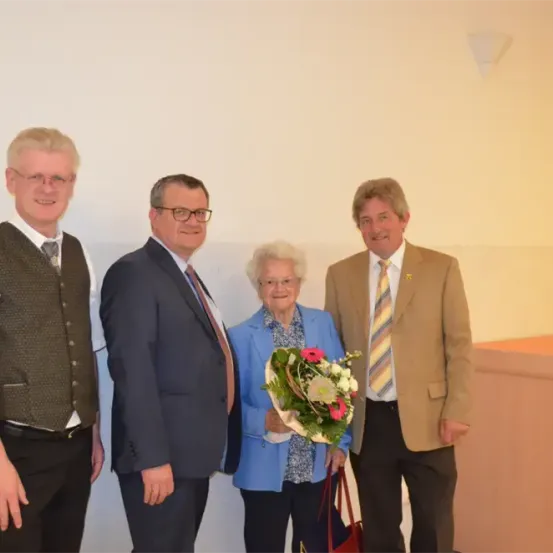 Vier Personen, zwei Männer und zwei Frauen, stehen lächelnd zusammen im Innenraum. Eine Frau hält einen Blumenstrauß.