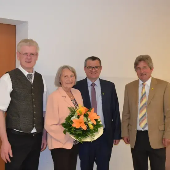 Vier Personen, die formell gekleidet sind, stehen nebeneinander, eine Frau hält einen Blumenstrauß. Sie scheinen für ein Foto in einem Raum zu posieren.