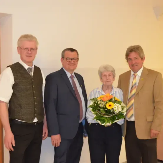 Vier Personen stehen zusammen und lächeln für ein Foto. Die Frau hält einen Blumenstrauß. Hinter ihnen ist eine schlichte Wand.