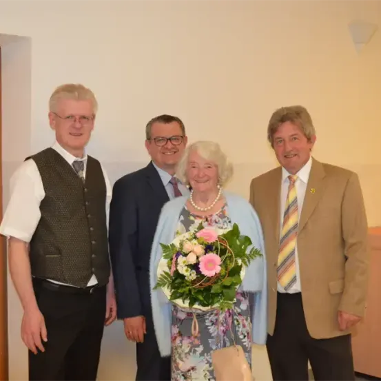 Vier Personen, darunter eine Frau mit einem Blumenstrauß, stehen lächelnd und feierlich gekleidet in einem Raum.